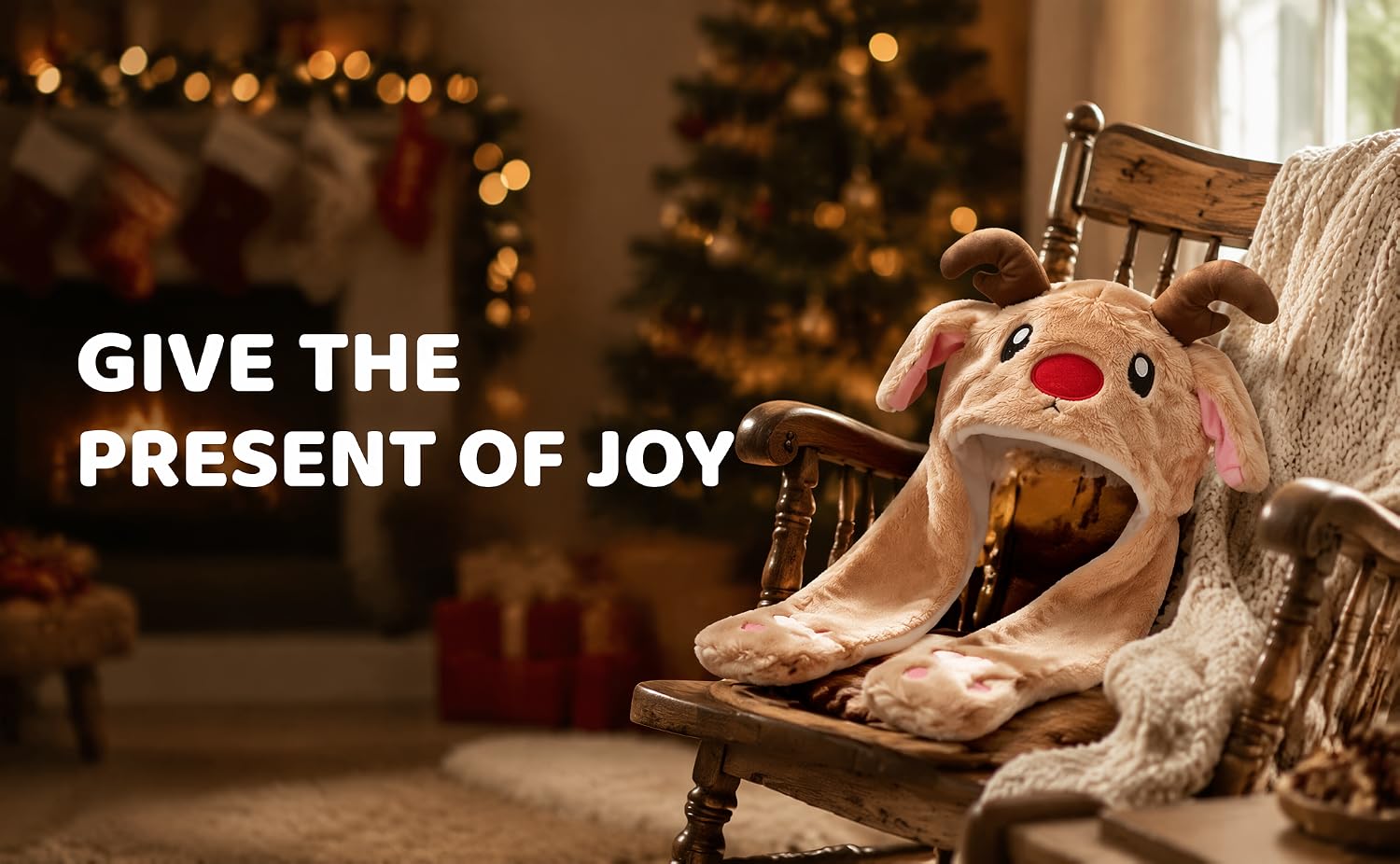 Text reads 'GIVE THE PRESENT OF JOY'. Reindeer plush toy with red nose sitting in wooden chair against festive Christmas background with blurred lights and decorations.
