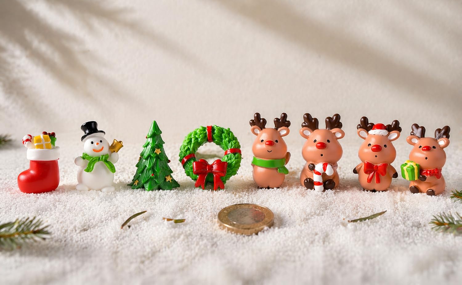 Collection of festive holiday decorative items including red ornaments, green wreaths, white snowflakes, and miniature Christmas character figurines arranged on white snowy surface.